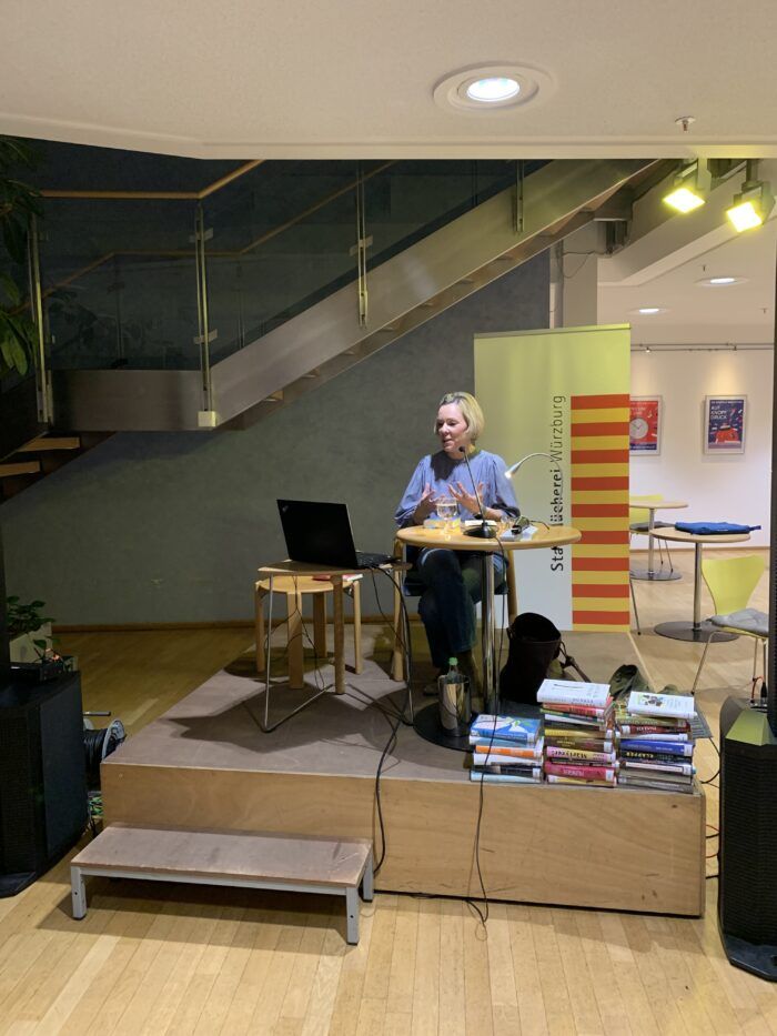 Auf diesem Foto ist die Referentin Frau Dr. Fraas bei der Veranstaltung zu sehen. Sie sitzt auf einer kleinen Bühne mit ihrem Laptop und vielen Büchern.