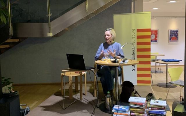 Auf diesem Foto ist die Referentin Frau Dr. Fraas bei der Veranstaltung zu sehen. Sie sitzt auf einer kleinen Bühne mit ihrem Laptop und vielen Büchern.